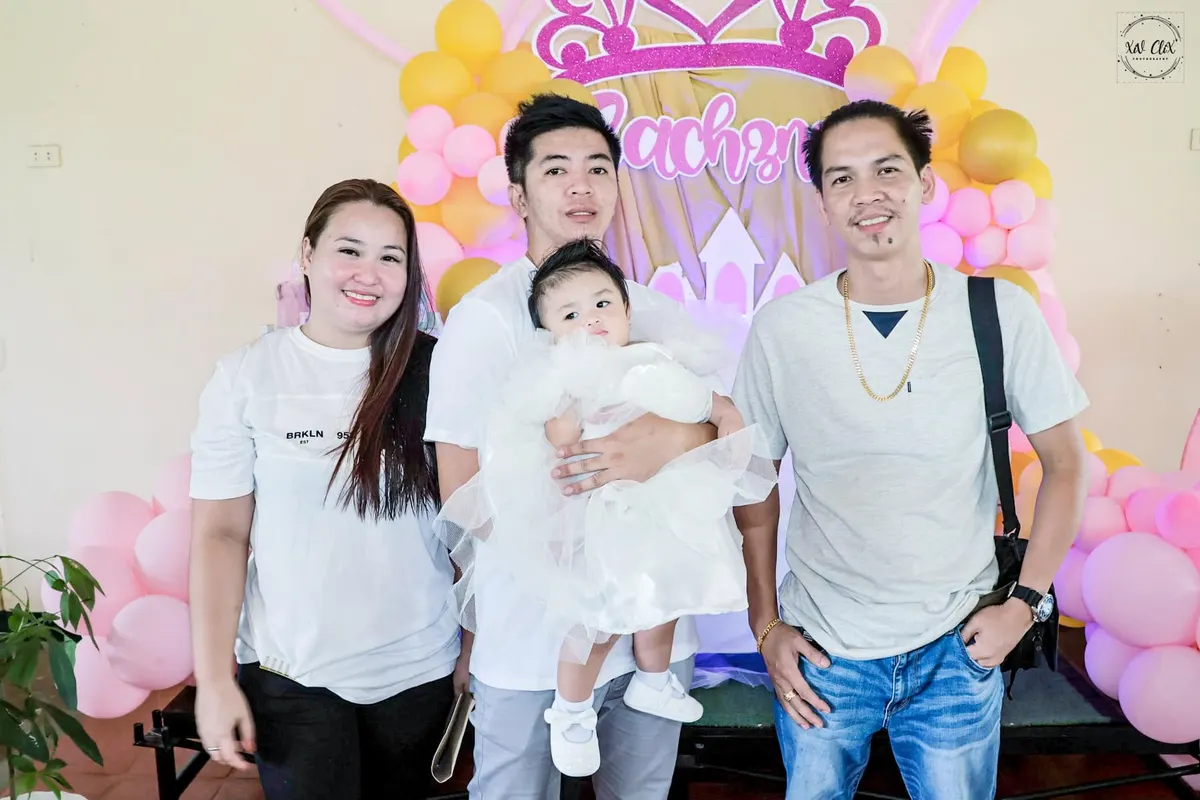Parents holding their baby in front of a pink balloon backdrop at a baptism reception