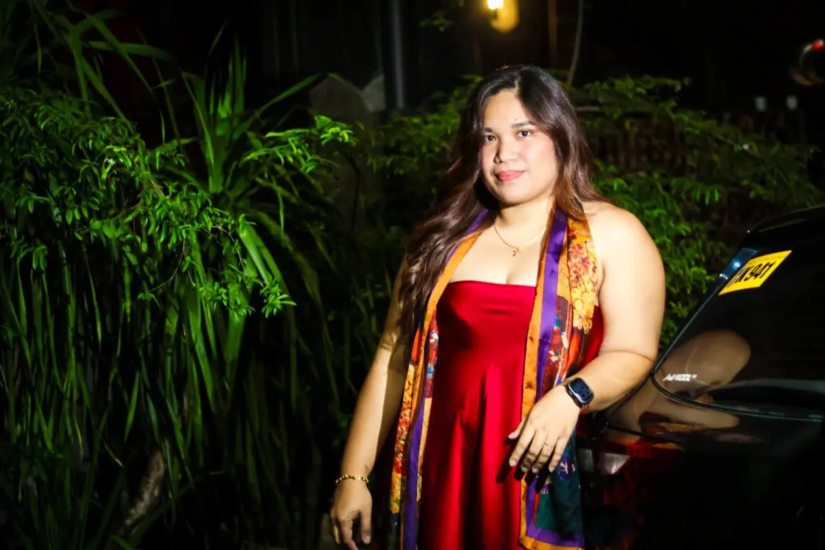 Woman in a red dress and colourful scarf among plants, lit at night