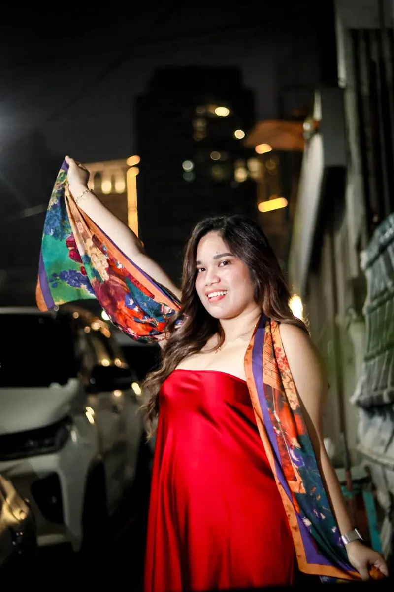 Woman in a red dress holding a colourful scarf beside parked cars at night
