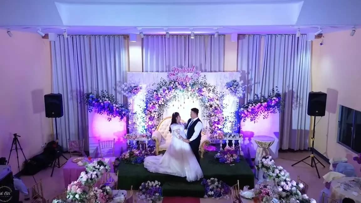 Couple on a flower-filled reception stage lit in lavender and blue
