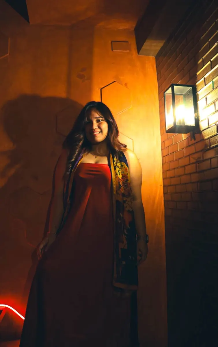Woman in a red dress standing beside a wall lamp in warm orange light