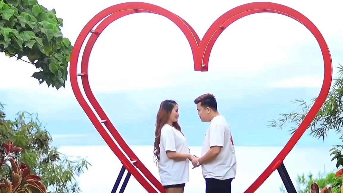 Couple standing inside a large red heart frame overlooking the sea